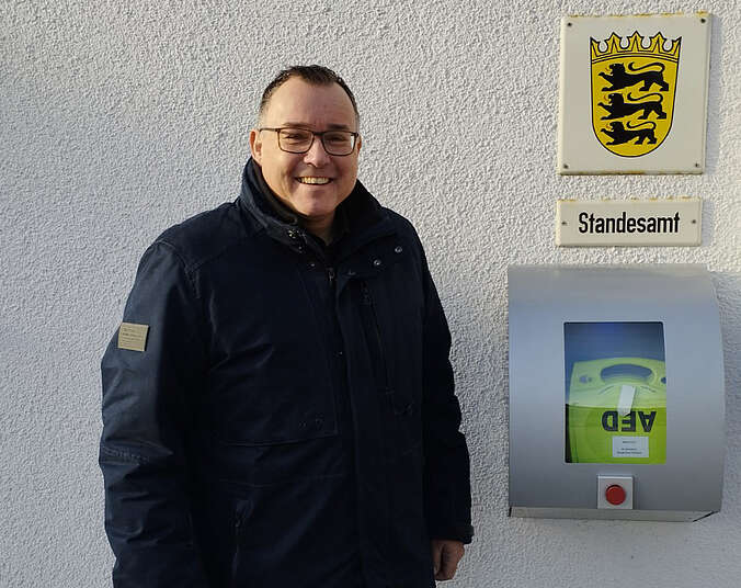 Mann steht neben Defibrillator darüber zwei HInweisschilder STandesamt und das Wappen Baden-Württemberg