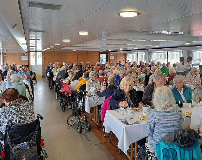 Männer und Frauen sitzen an Tischen im Innenbereich des Schiffes, trinken Kaffee und essen Kuchen.